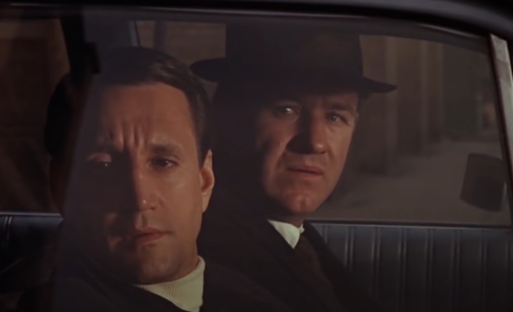 Gene Hackman in The French Connection, in suit and a hat sitting in a police car