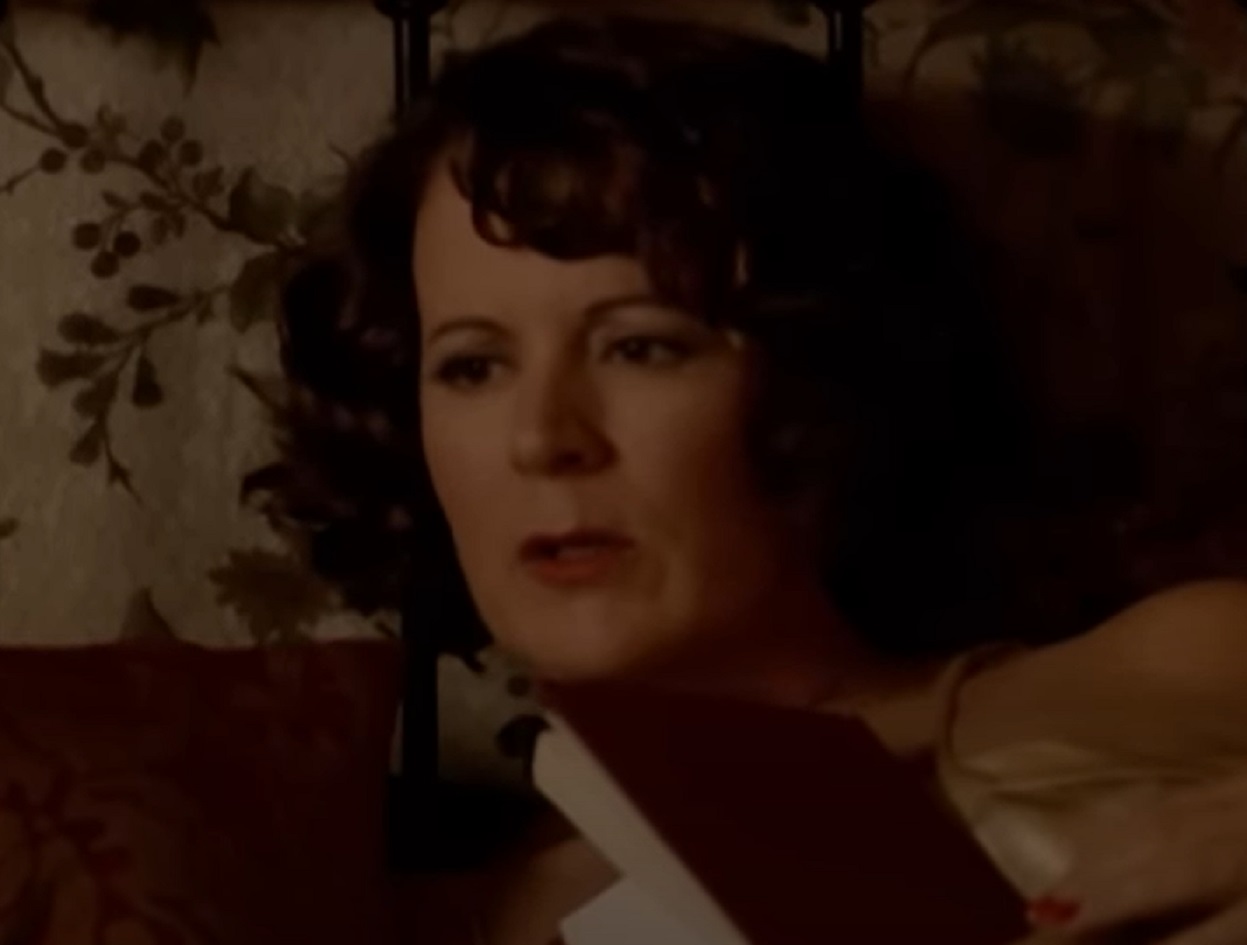 Screenshot: Gladys Pearl Baker (Patricia Richardson) is laying on the bed ,holding a book - from Blonde (2001)
