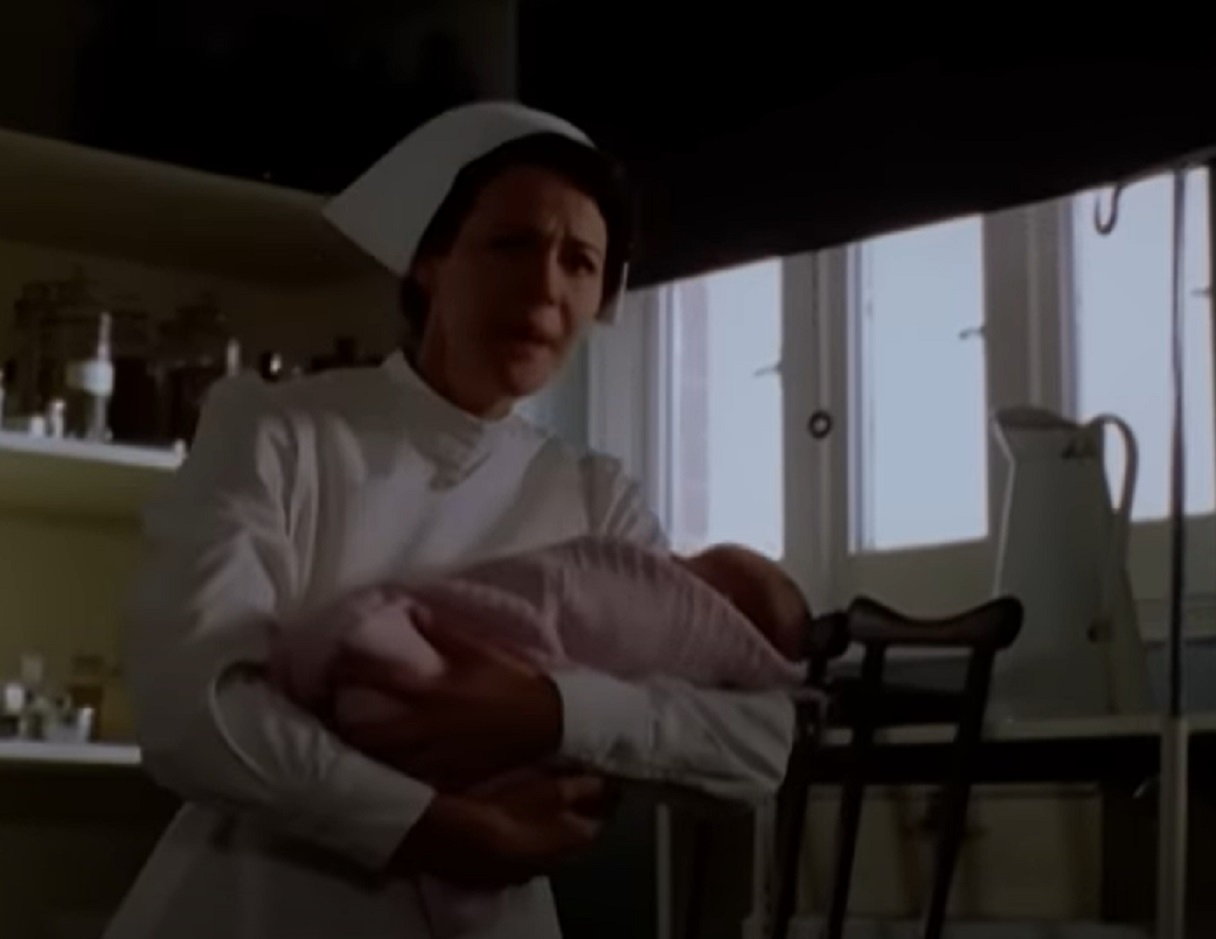 Screenshot: Young nurse in hospital is holding a small baby and talking at camera - from Blonde (2001)