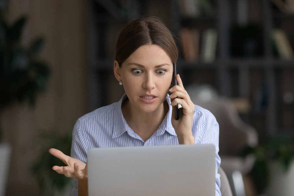 Shocked woman speaking on phone and working on laptop in office