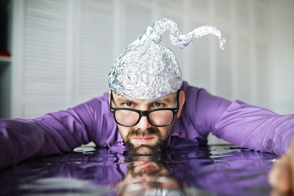 Bearded funny man wearing cap of aluminum foil believing in conspiracy theories in purple shirt
