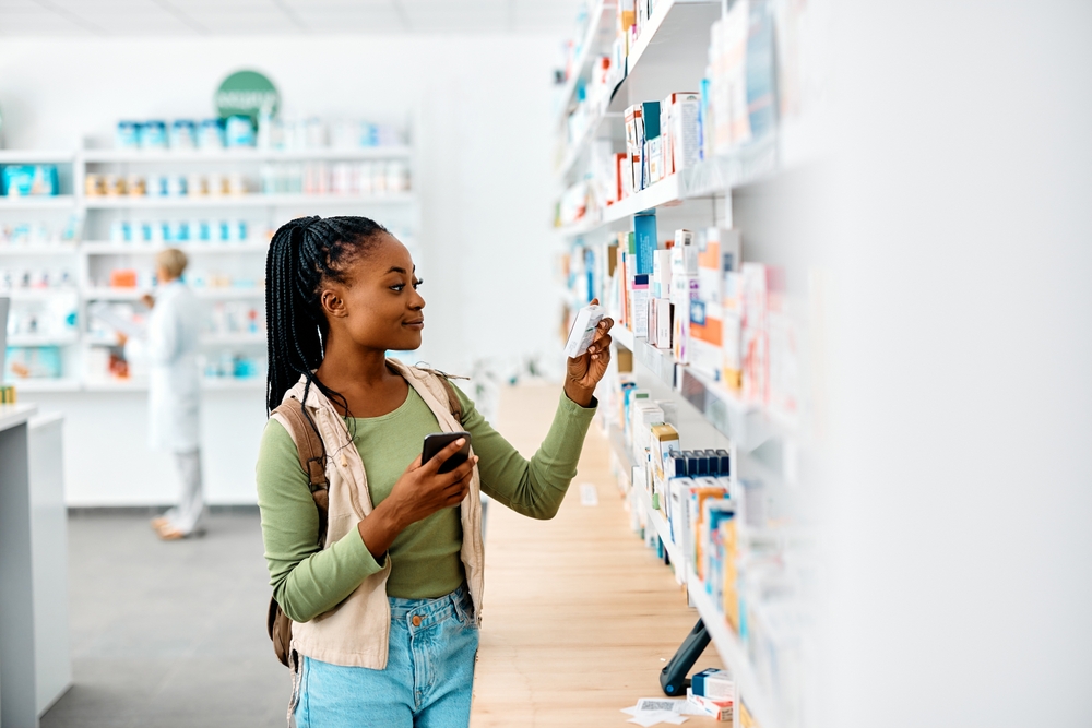 Young African American woman using cell phone while looking for a medicine in pharmacy wearing green top