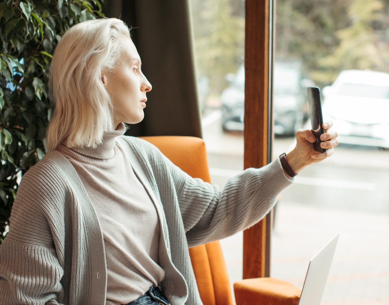 Blonde woman is seating in room and making a selfie with her phone.