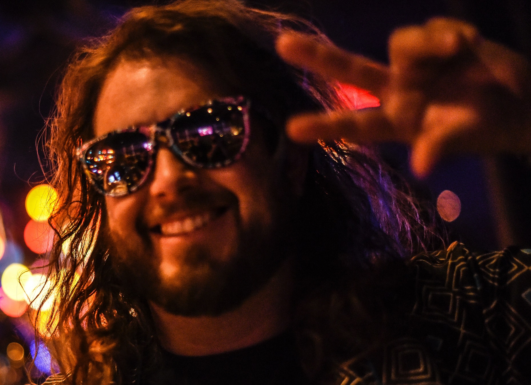 Man with long hair and dark glasses is smiling and making a peace sign.