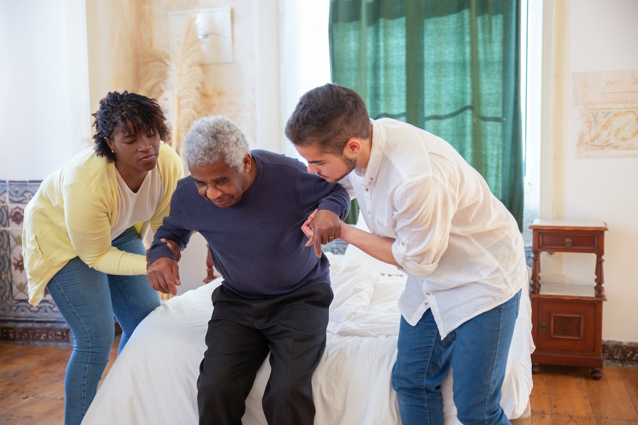 A man and a woman assisting an elderly man in standing from the bed.