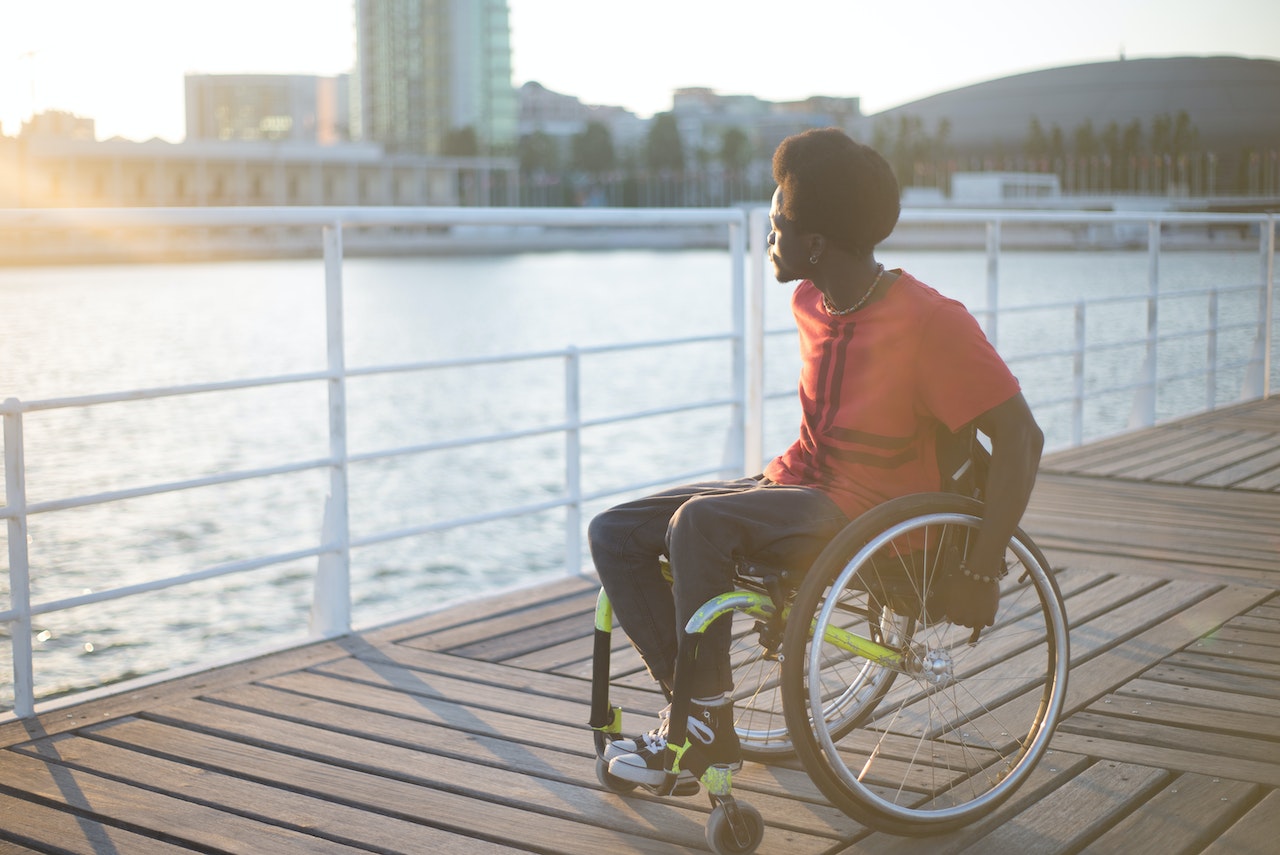 Young black man is seating at wheelchair and looking at water outside.