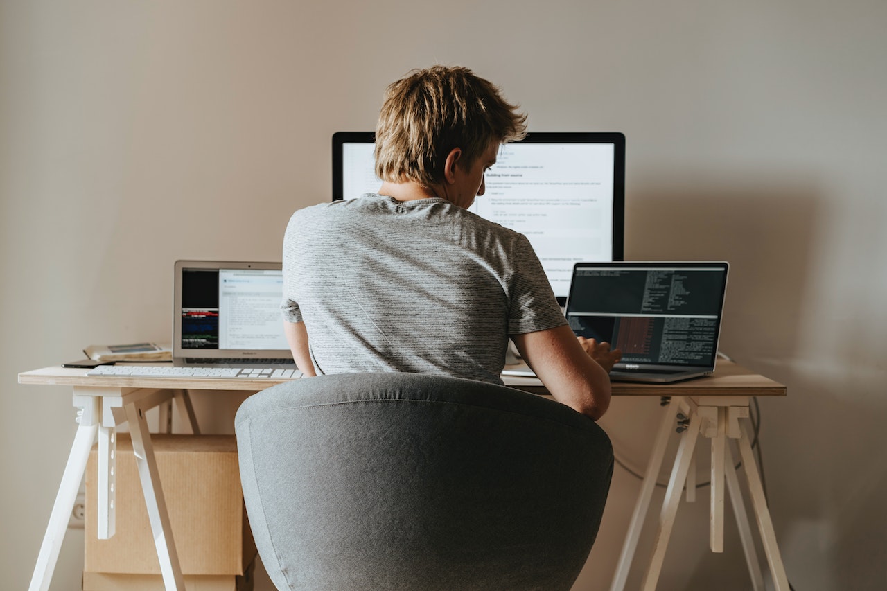 Young man in grey shirt is seating on desk and coding on a PC.