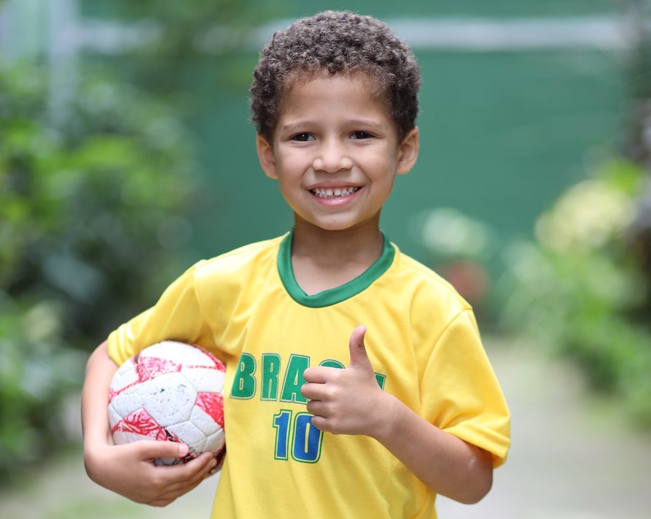 Small kid wearing yellow shirt is holding a ball and smiling at camera.