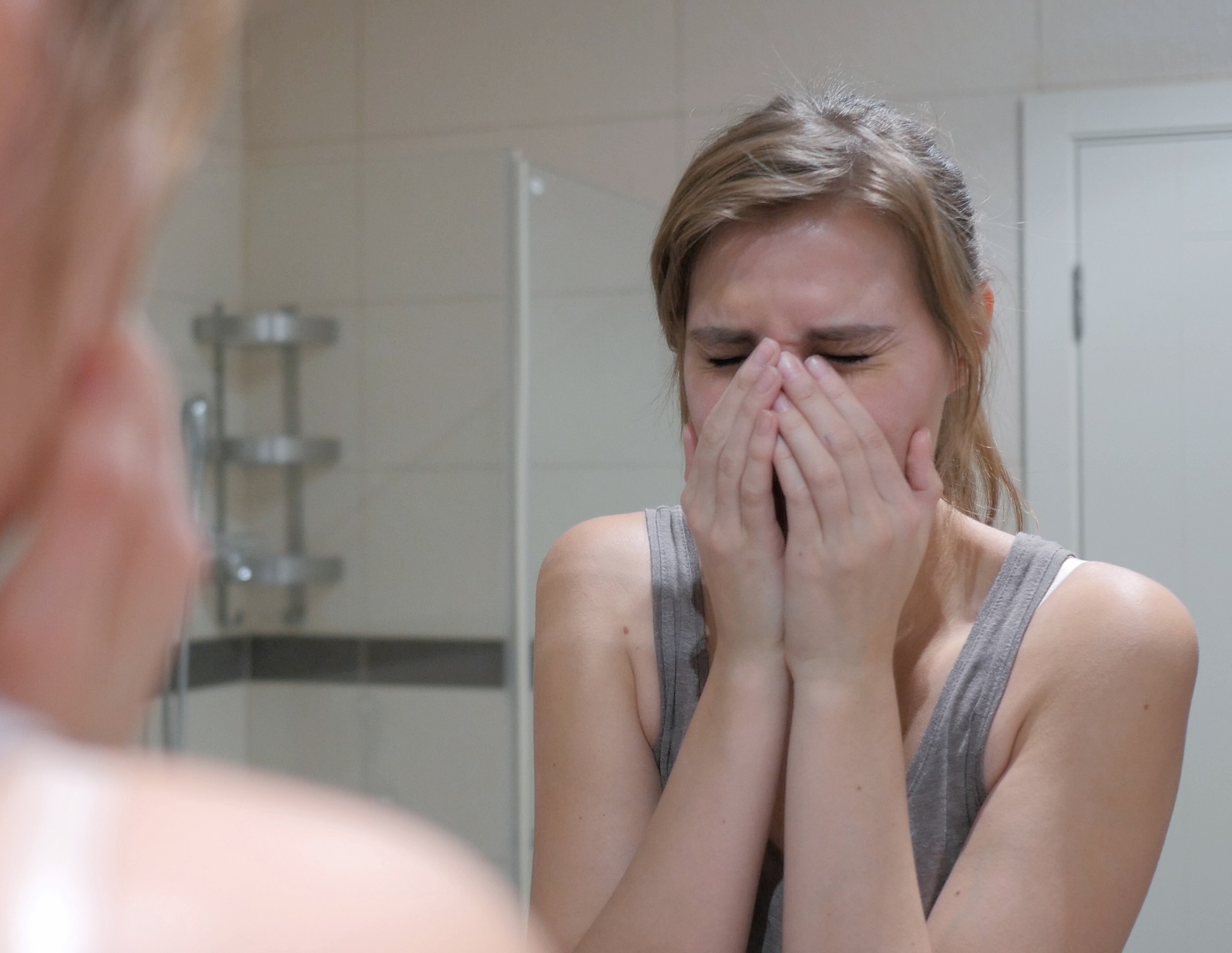 Woman is standing in front of a mirror in bathroom and crying.