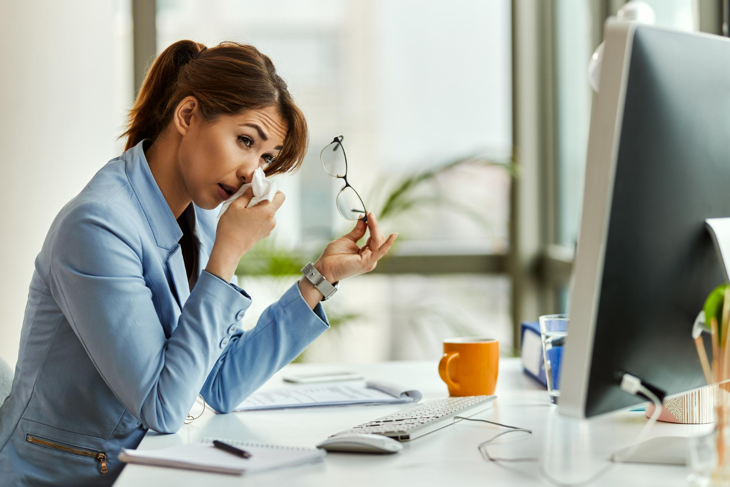 Woman in blue jacket is seating at her office desk and crying.