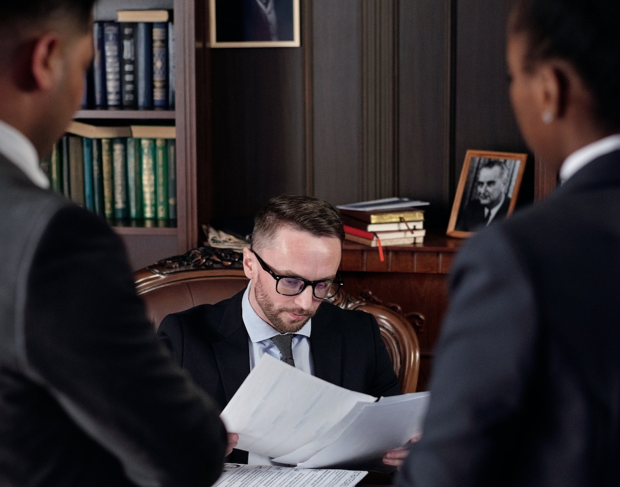 Two lawyers is standing next to a man looking in to documents seating at desk in office.
