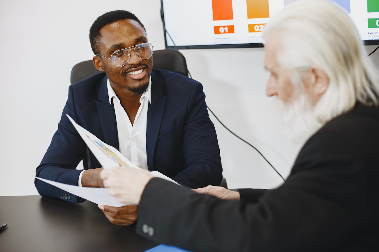 Senior man with white hair and beard is talking with young black man who is smiling at office.