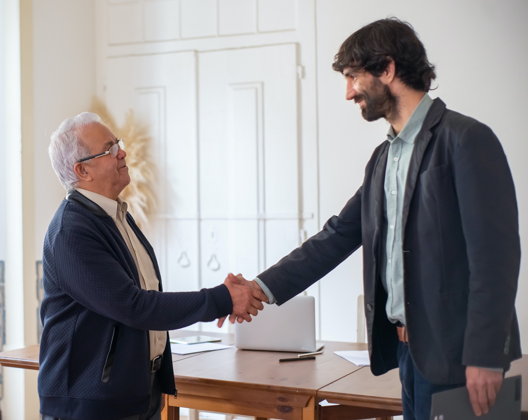 Senior man is handshaking with young bearded man at office.