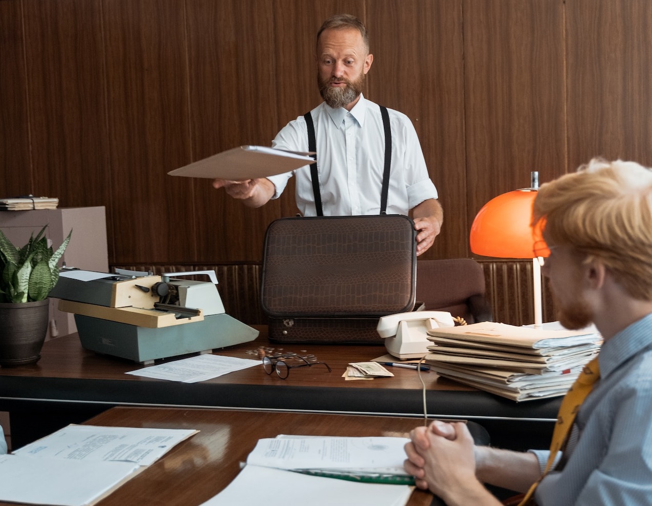 Man is holding a papers in his hand and talking with man seating on his desk at office.