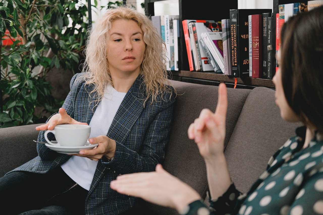 Woman is talking and pointing to a woman drinking coffee at office.