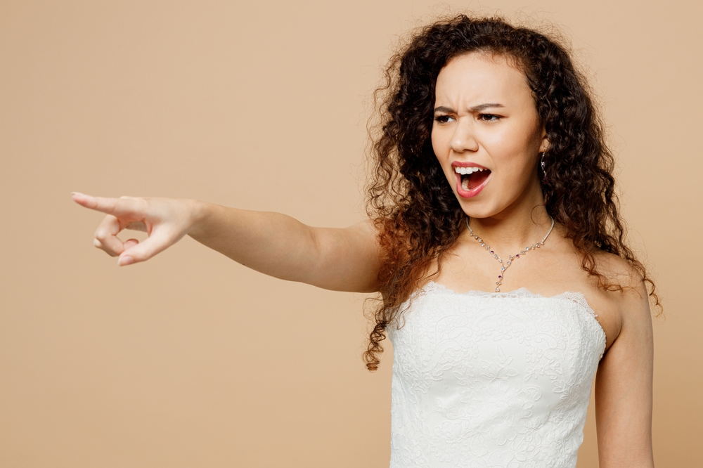Sad young angry bride wearing white wedding dress pointing with her index finger aside