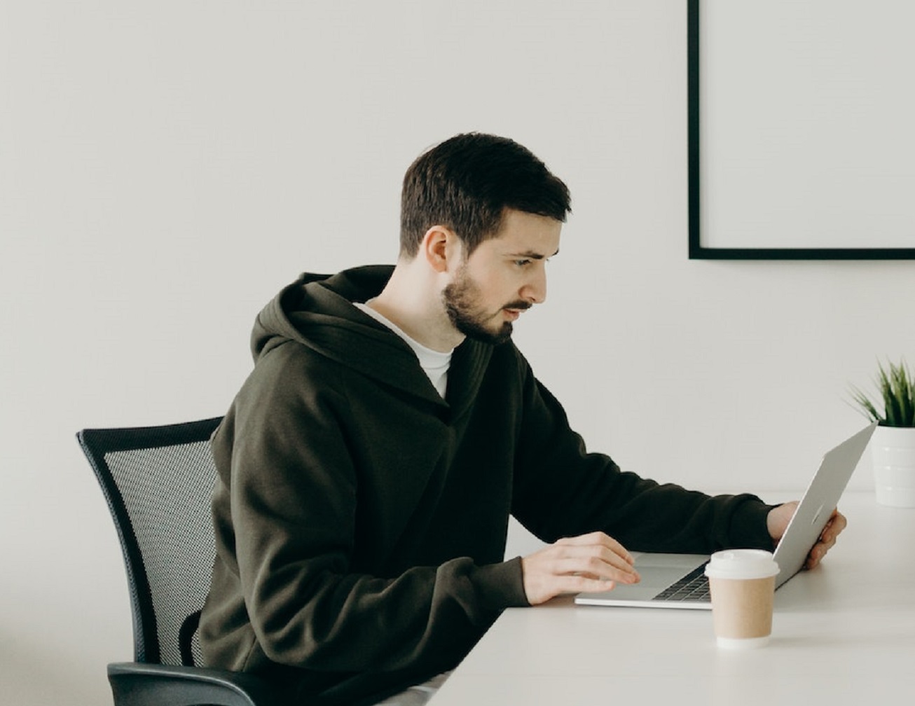 Young bearded man is working on laptop on his desk.