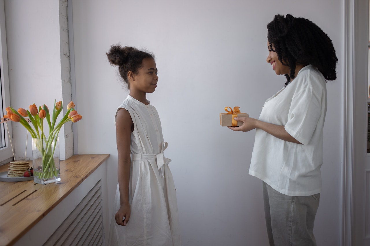 Young black woman is giving a present to a black girl wearing white dress in the room.