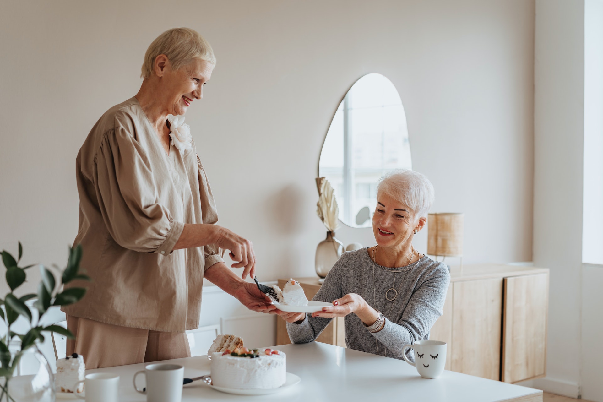 Senior woman is cutting a white cake on the kitchen table.