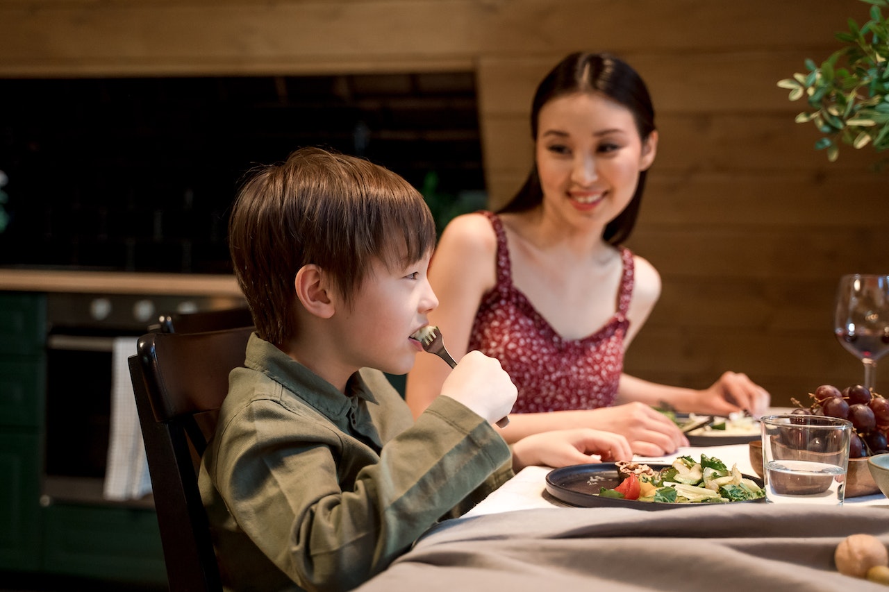 Young woman is having a dinner with her small son, eating food from the table.