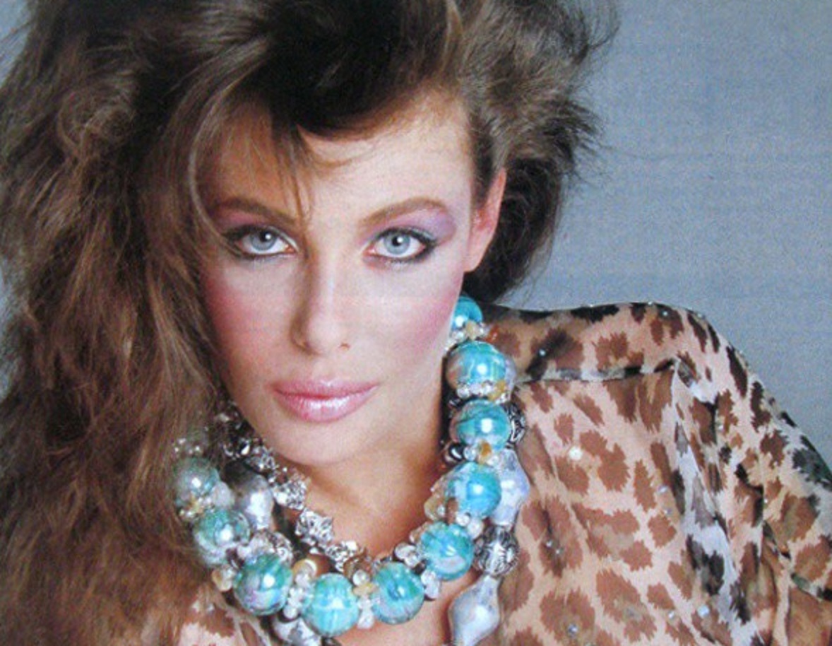 Kelly LeBrock wearing big blue necklace looking at camera.