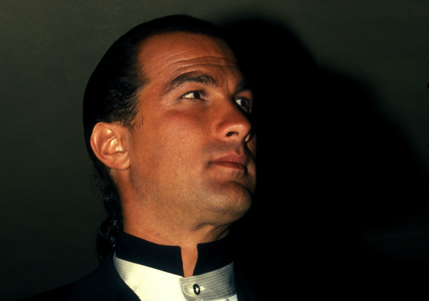Steven Seagal wearing black jacket is looking at side in dark room - 1991