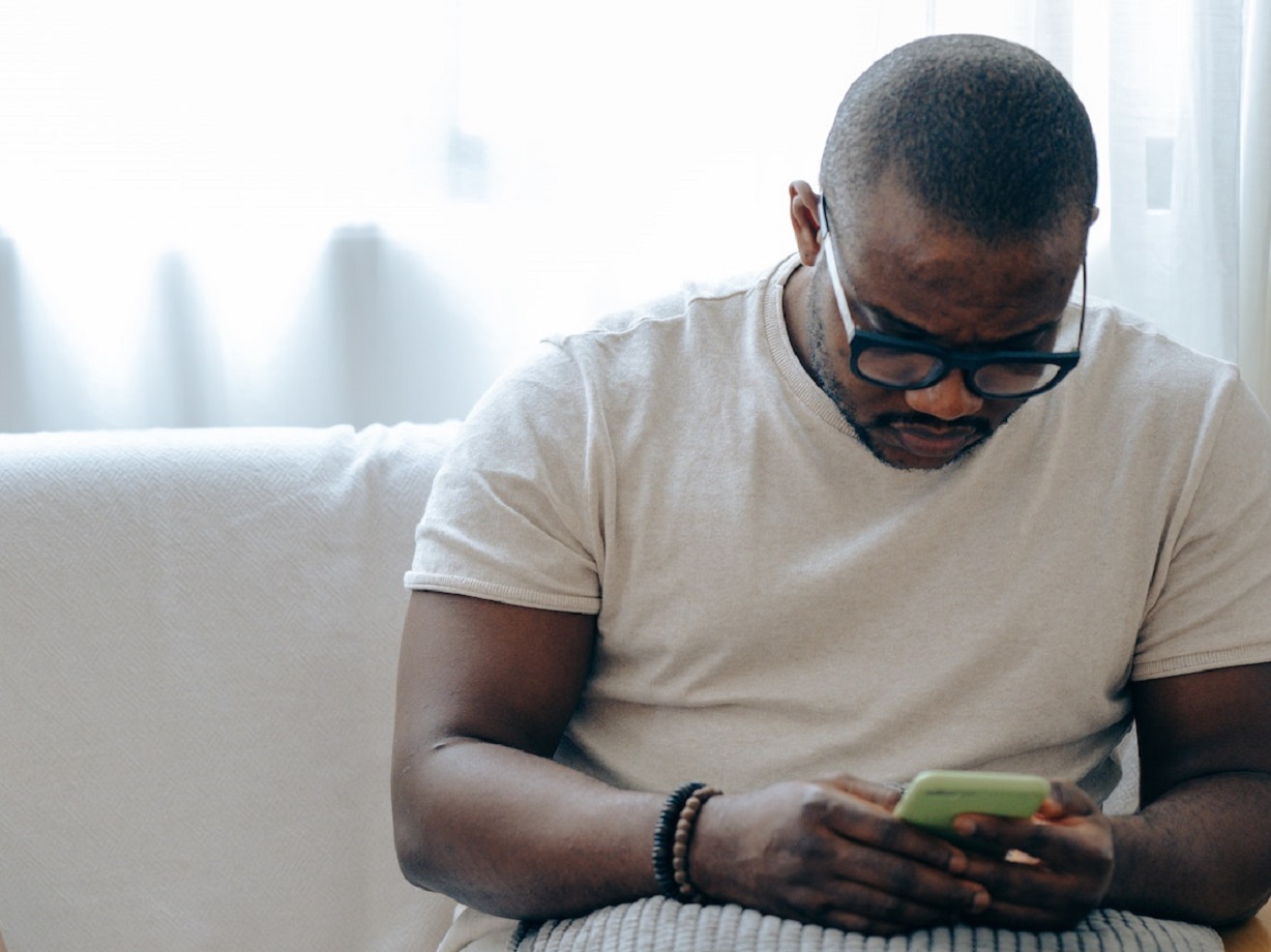 Man is seating on the bed and texting on his phone.
