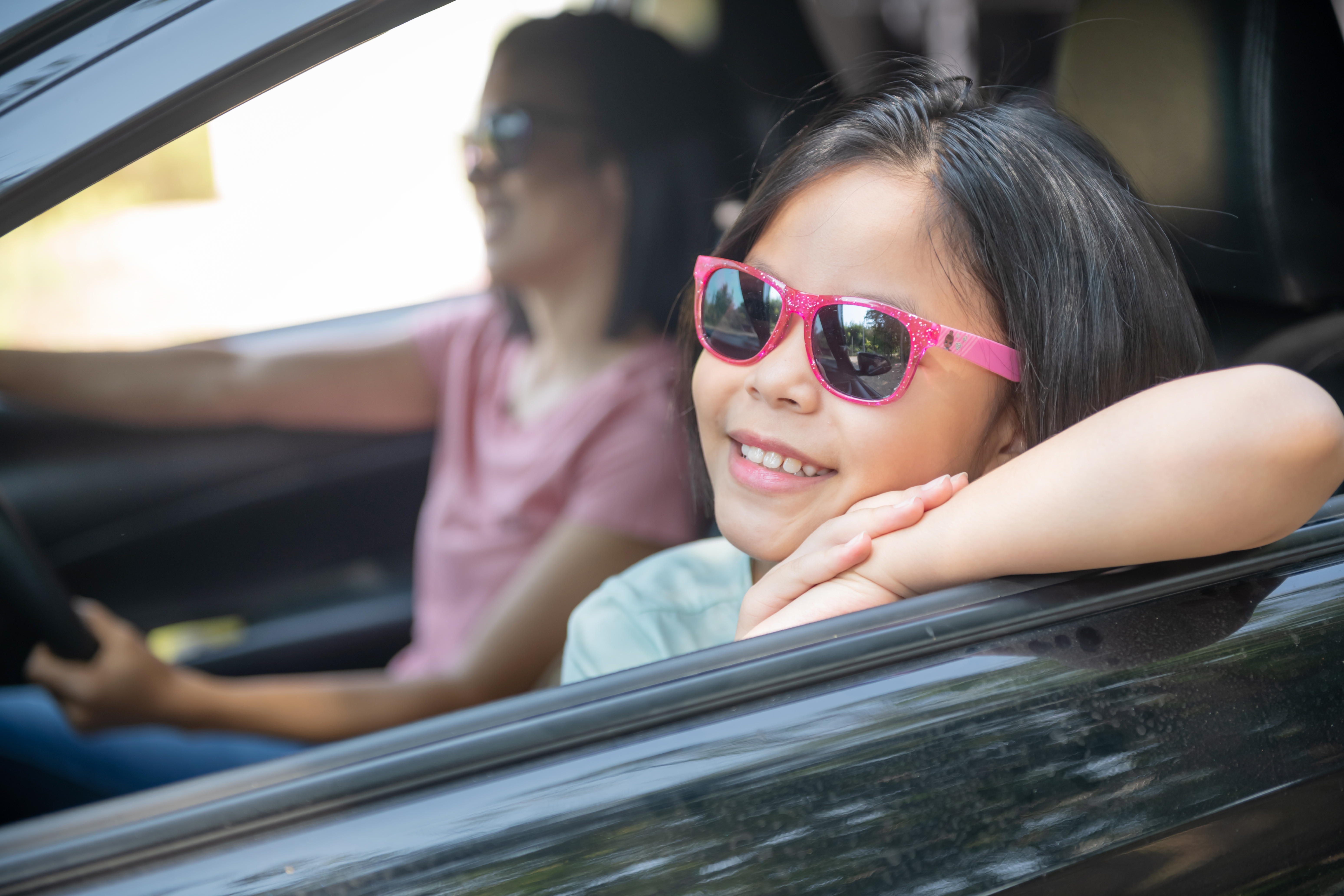 Girl wearing pink glasses and smiling is seating in the car with her mother.