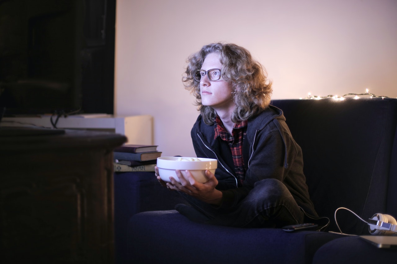 Young man is seating in front of a TV and eating popcorn.