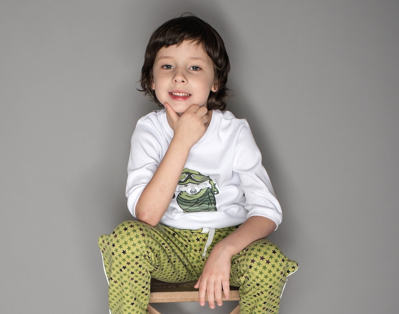 Small kid is seating on the chair and looking at camera.