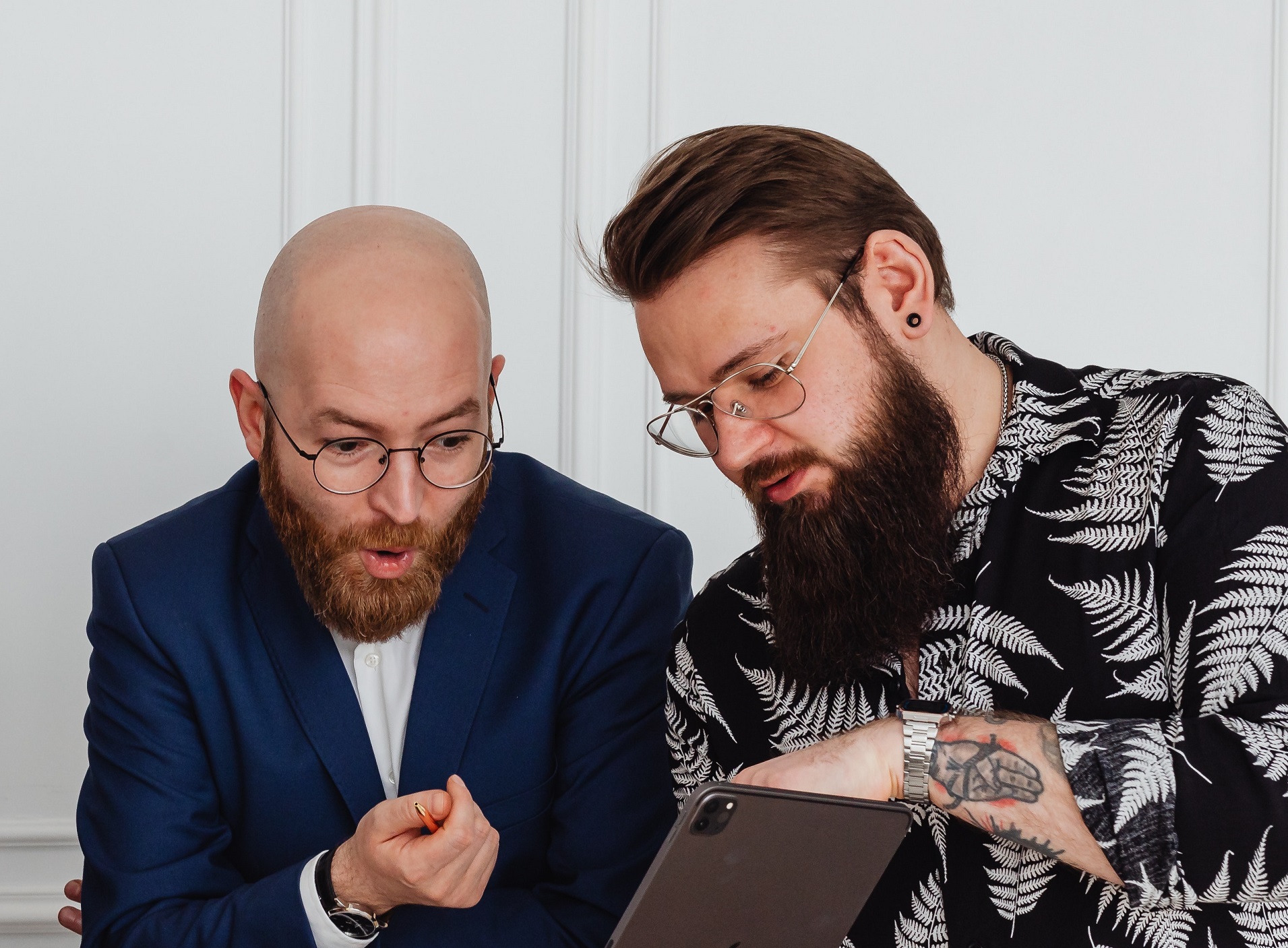 Bold bearded man is standing next to a man and looking surprised at tablet.