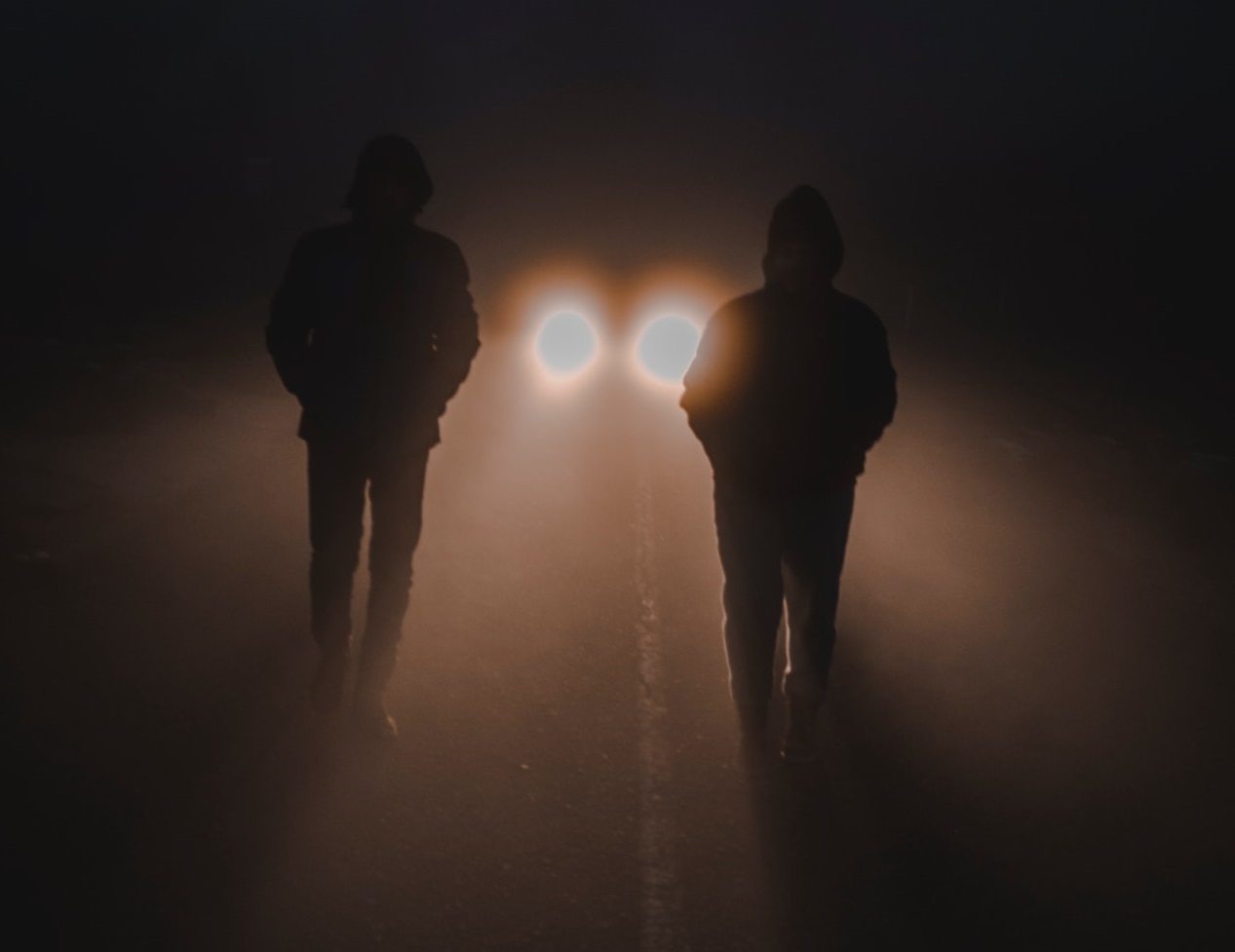 Two people are walking on a empty road at night with car light behind him.