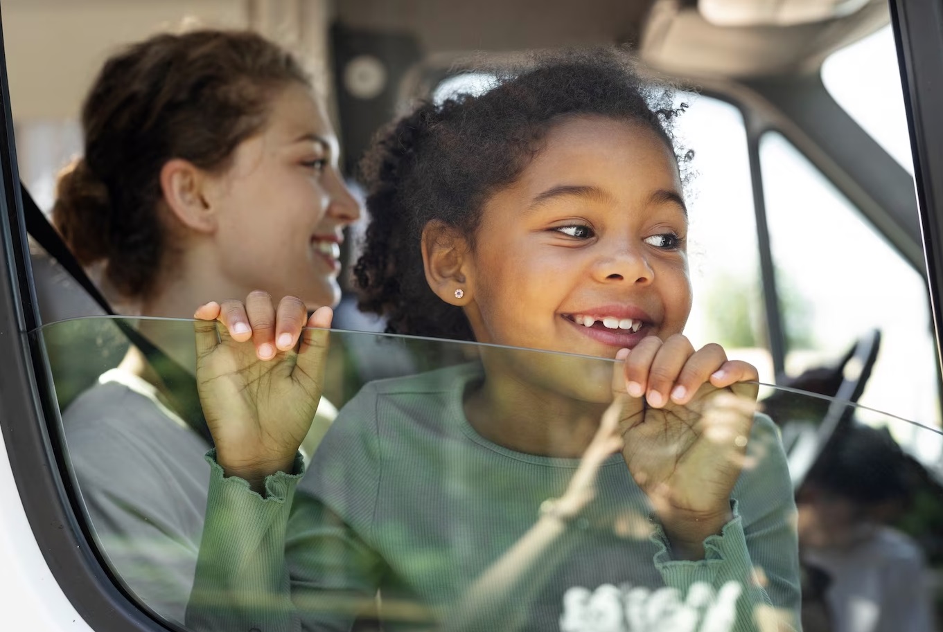 Young girl in grey shirt is smiling and looking thru window of a car.