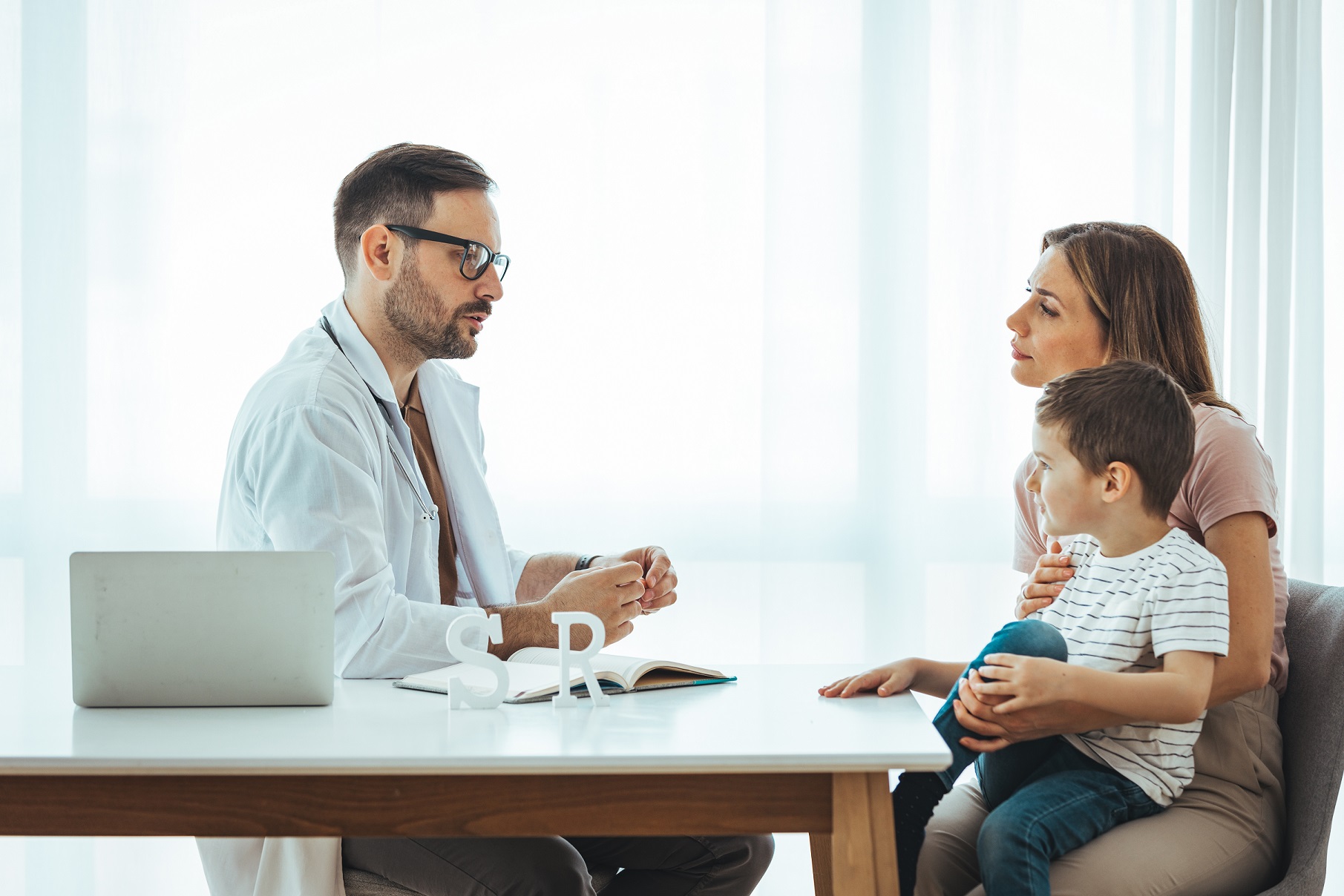 Mother holding her young son is talking with a young male doctor at his office.