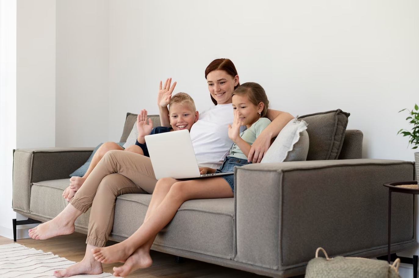 Woman seating with two kids on the sofa and having a video call on laptop.