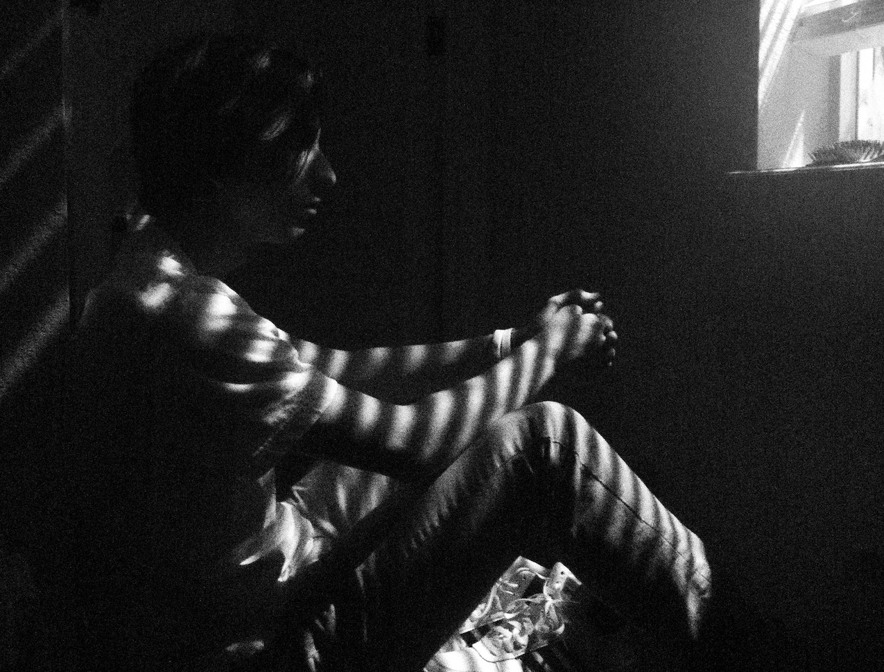 Sad man is seating next to his bed in dark room and thinking.