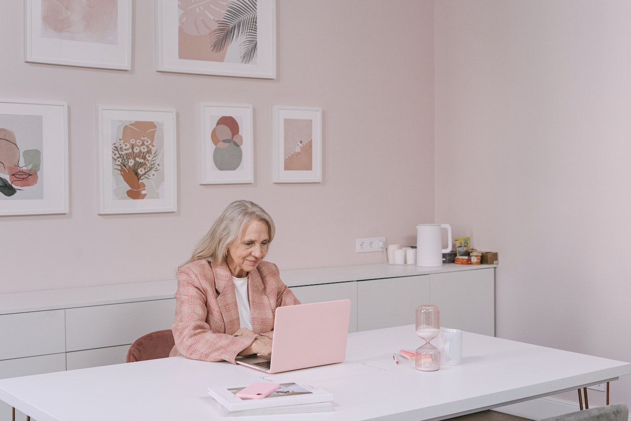 Senior woman is seating on desk and working at pink office.