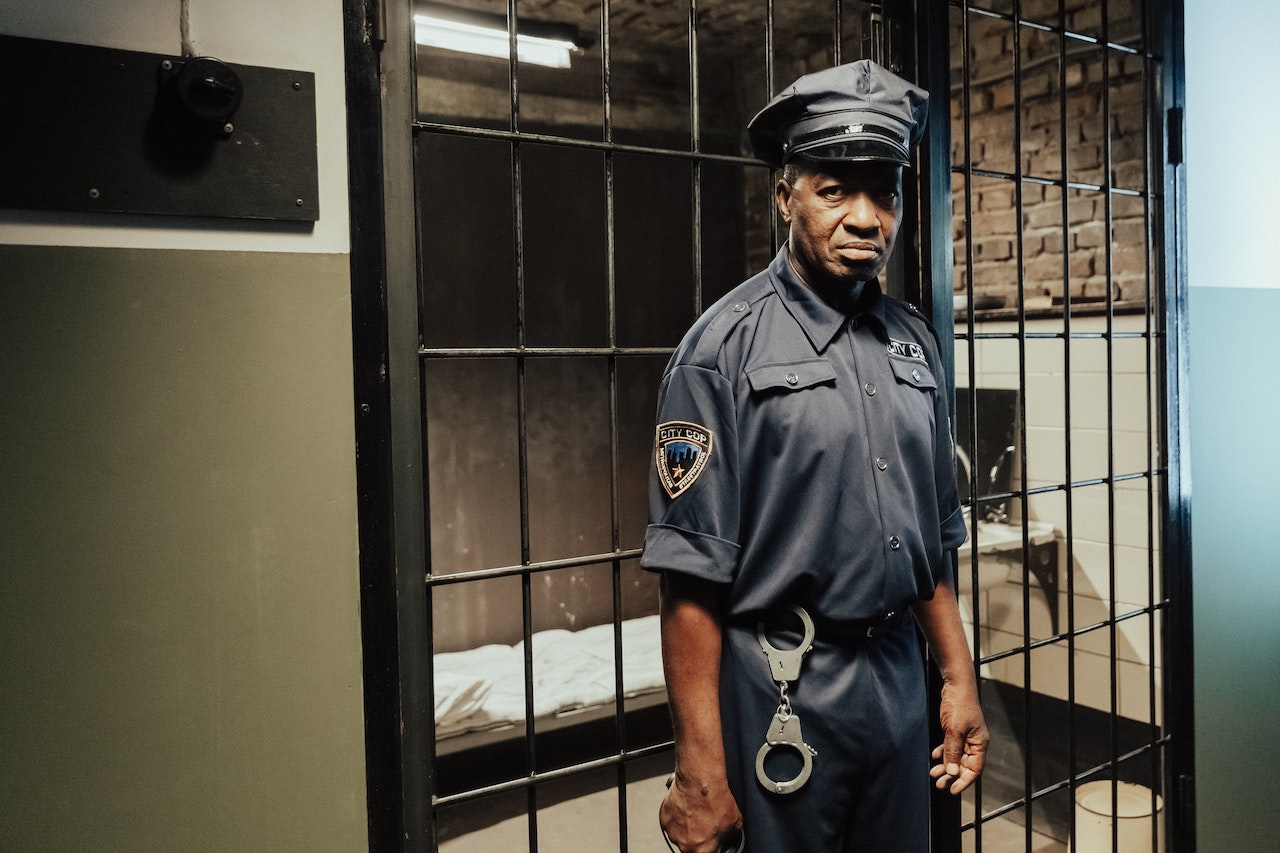 A black prison guard is standing by a empty prison cell.