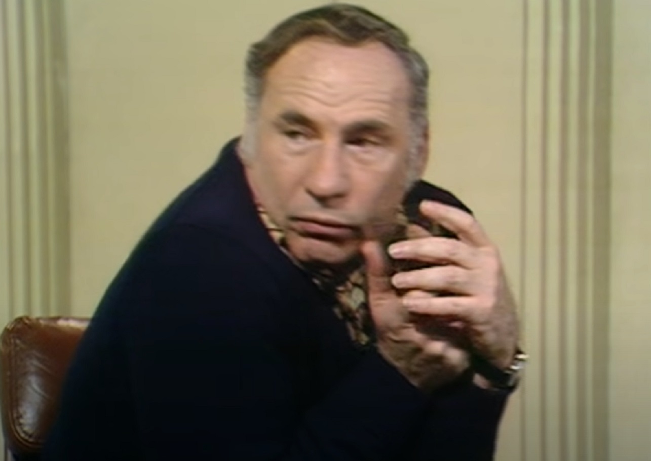 Screenshot: Mel Brooks wearing black suit is seating on chair and looking at side - from Mel Brooks-Unwrapped (2018)