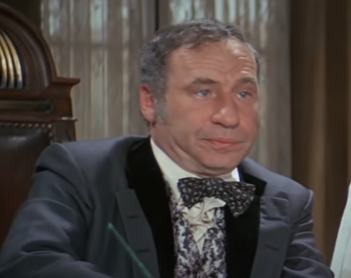 Screenshot: Mel Brooks wearing grey suit and bow tie looking at side - from Blazing Saddles (1974)