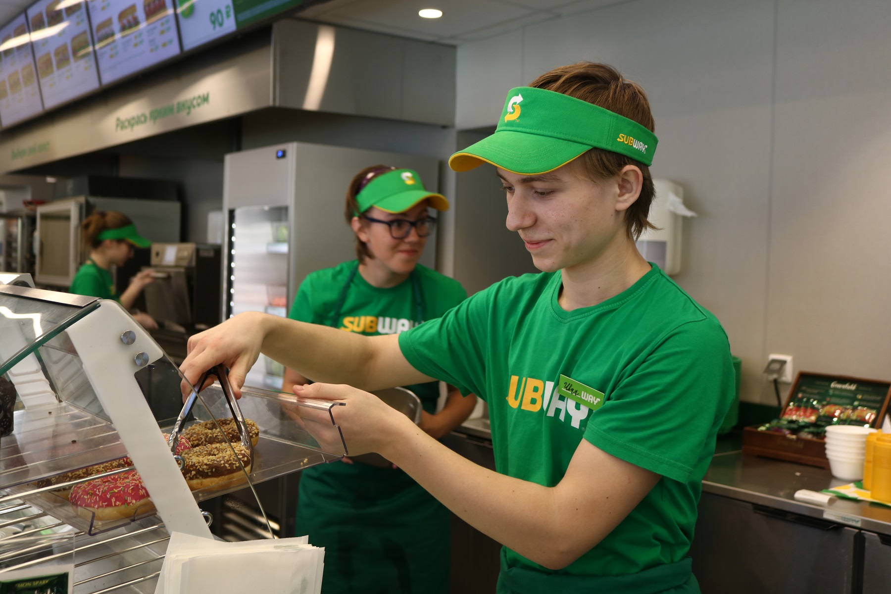 Subway worker wearing green uniform is serving a food.