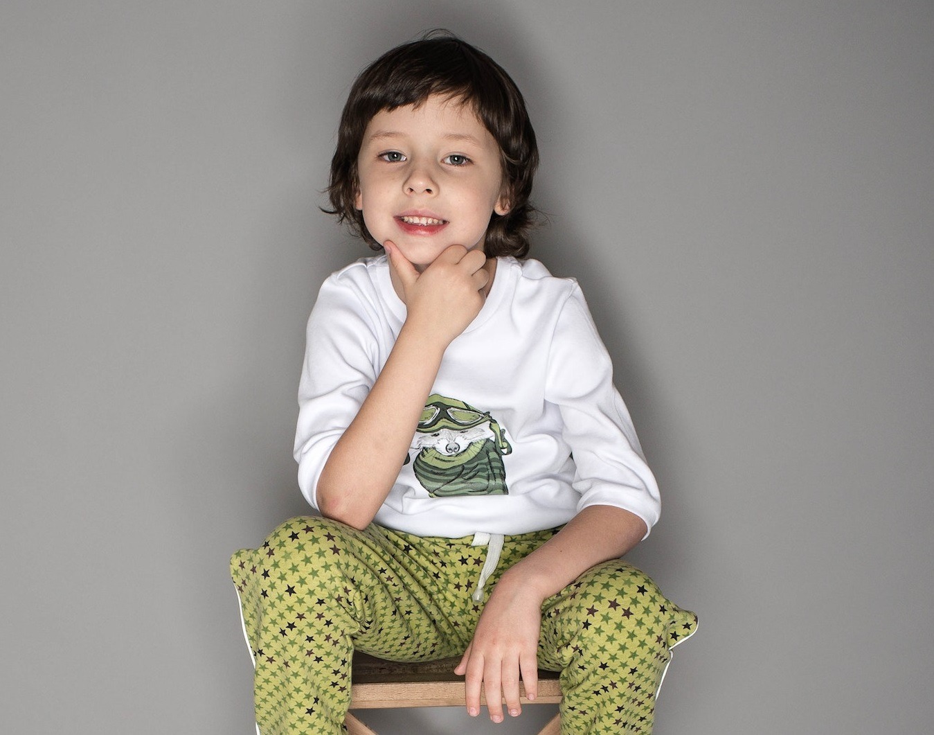 Small kid is seating on the chair and looking at camera.