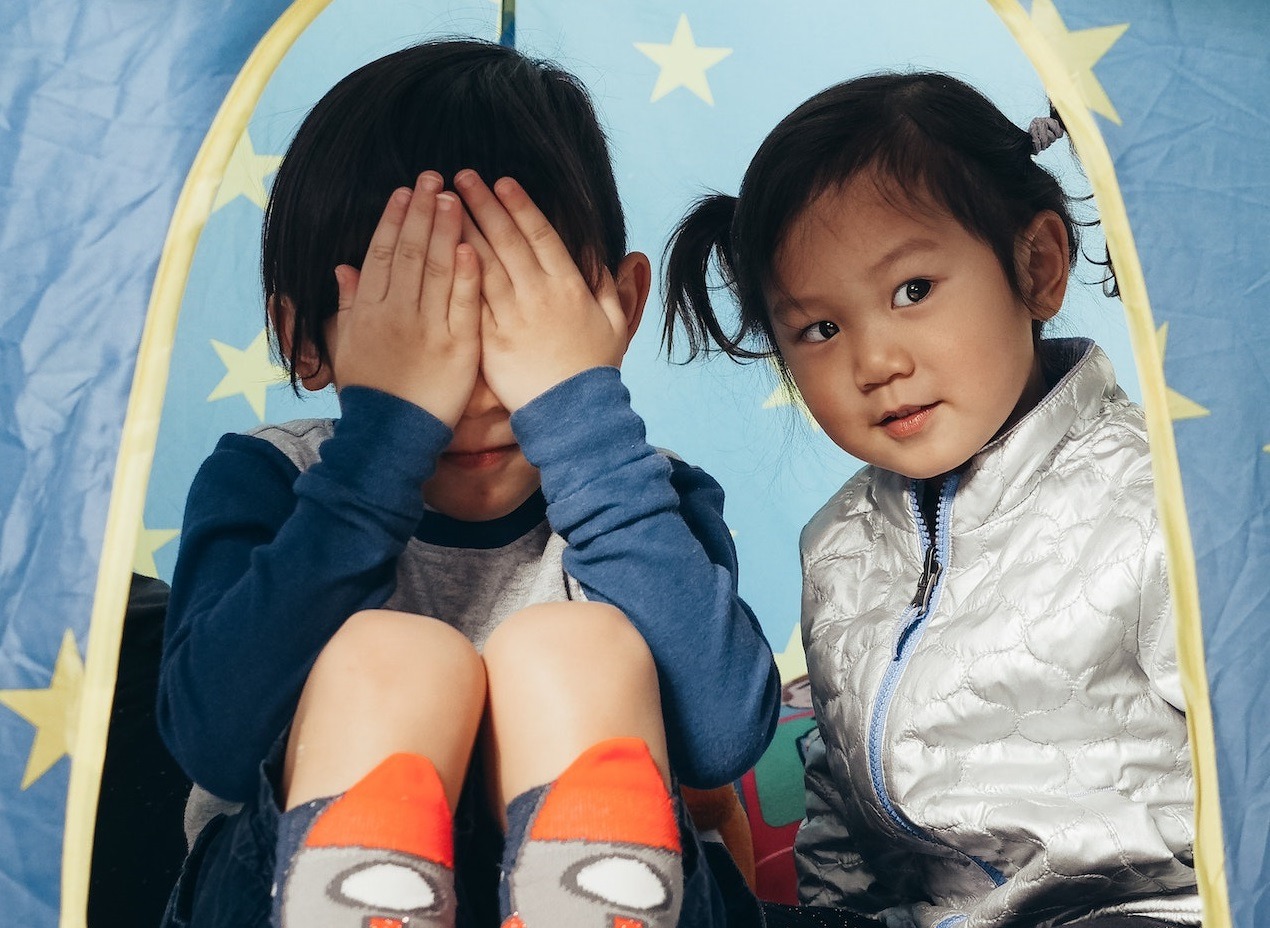 Small boy is covering his eyes and girl looking at side are seating in tent and talking.