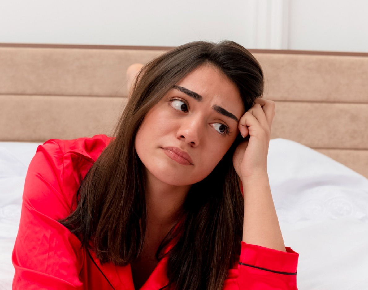 Young woman with long hair is seating in bed with upset face.