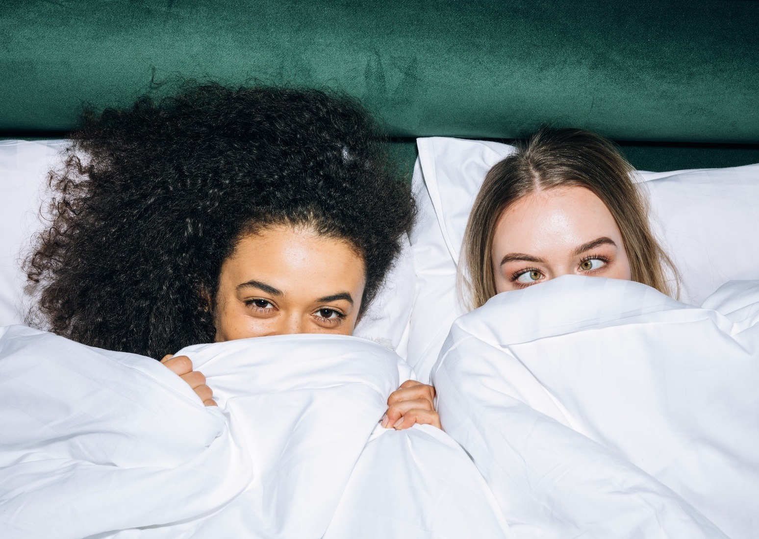 Two girls are hiding under a white sheet on the bed.