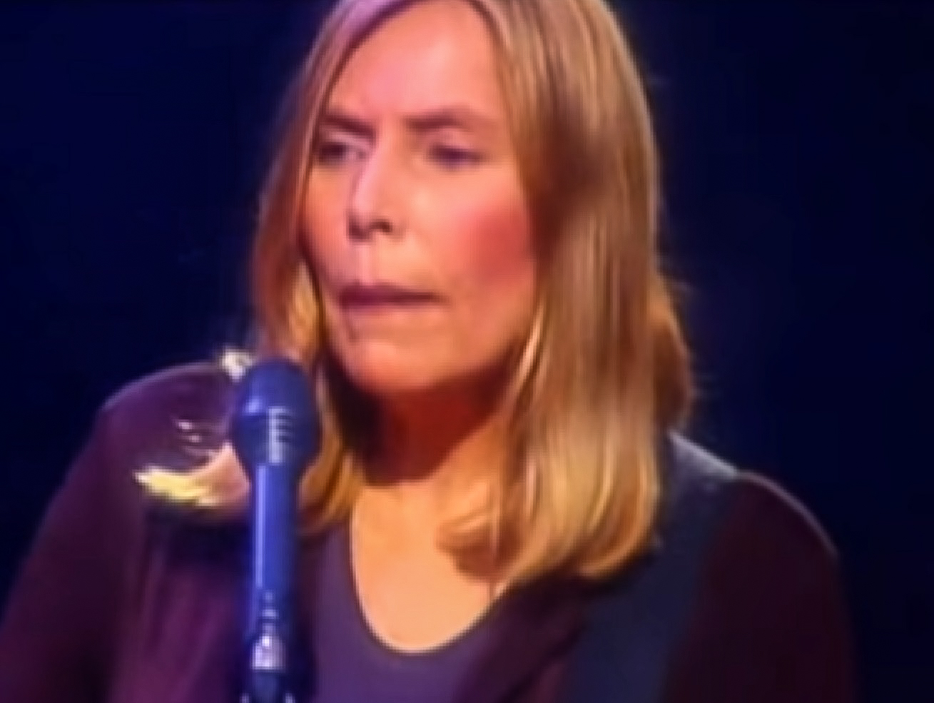 Screenshot: Joni Mitchell wearing black sweater is looking sad on stage - from Joni Mitchell: A Woman of Heart and Mind (2003).