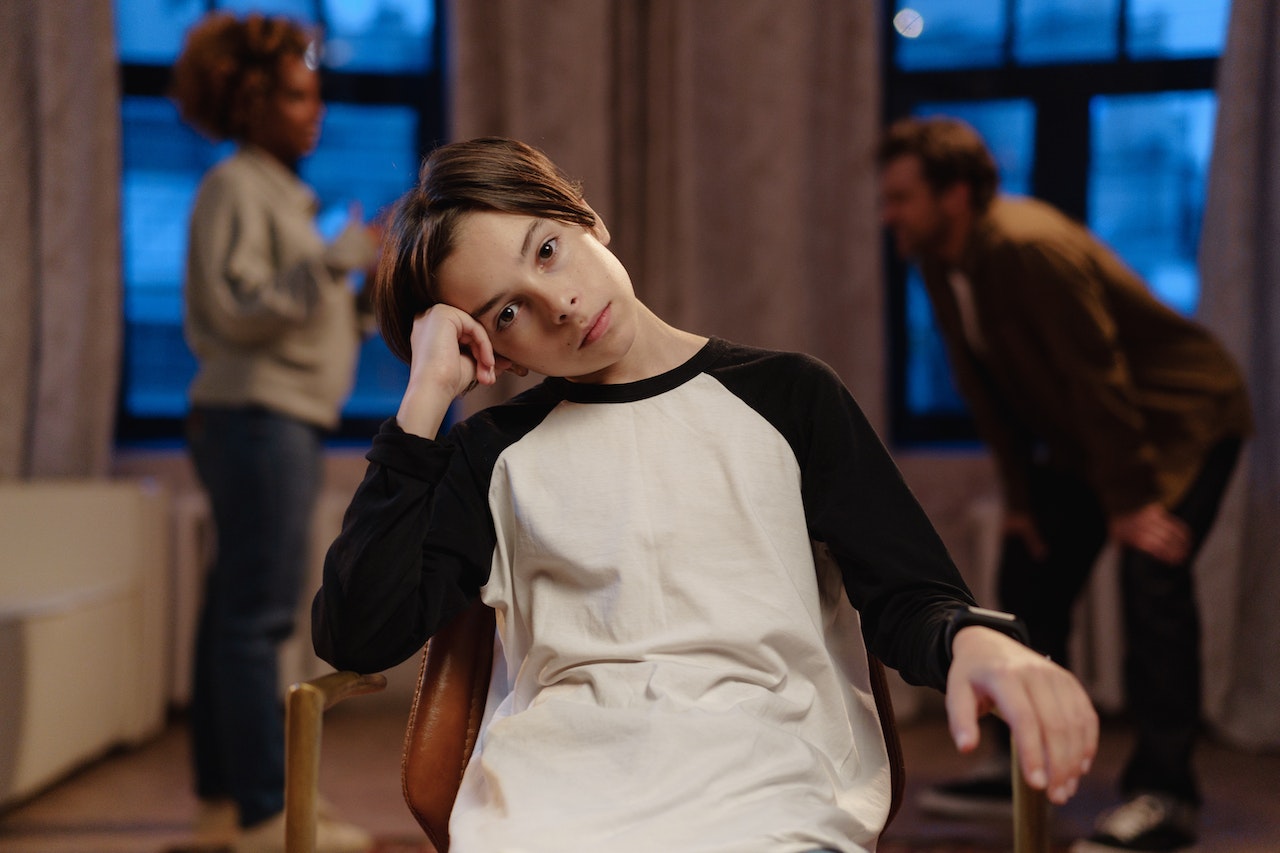 A boy is sitting on a chair while his parents are arguing behind him.