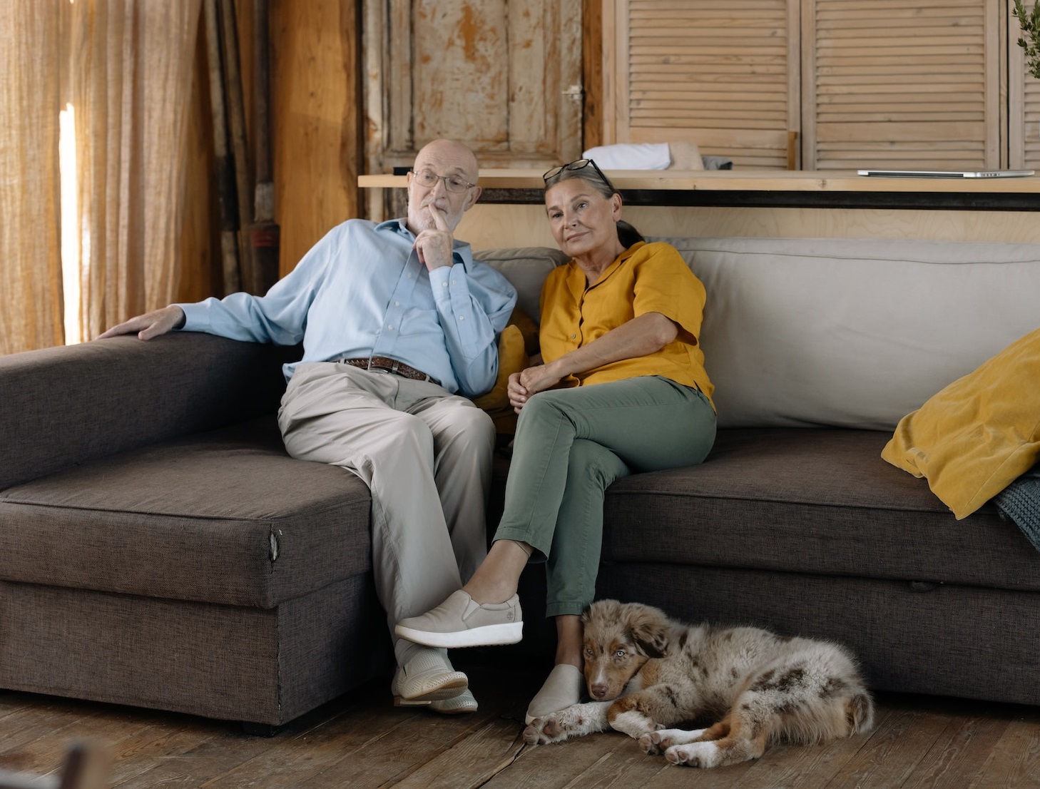 Senior couple is seating on the bed and looking at front with dog laying next to them.