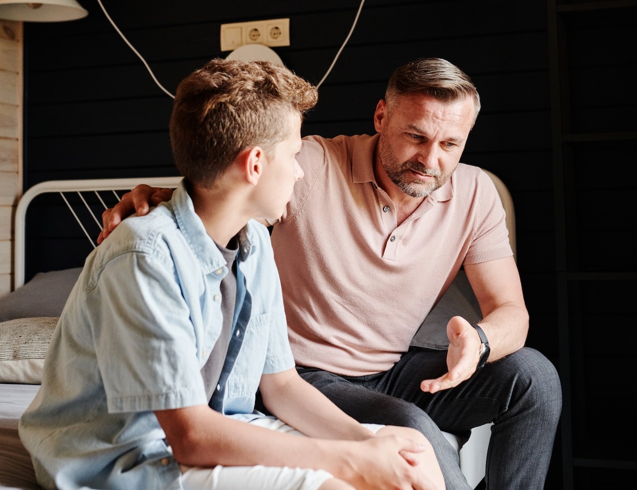 Father with beard is talking with his son seating on the bed.