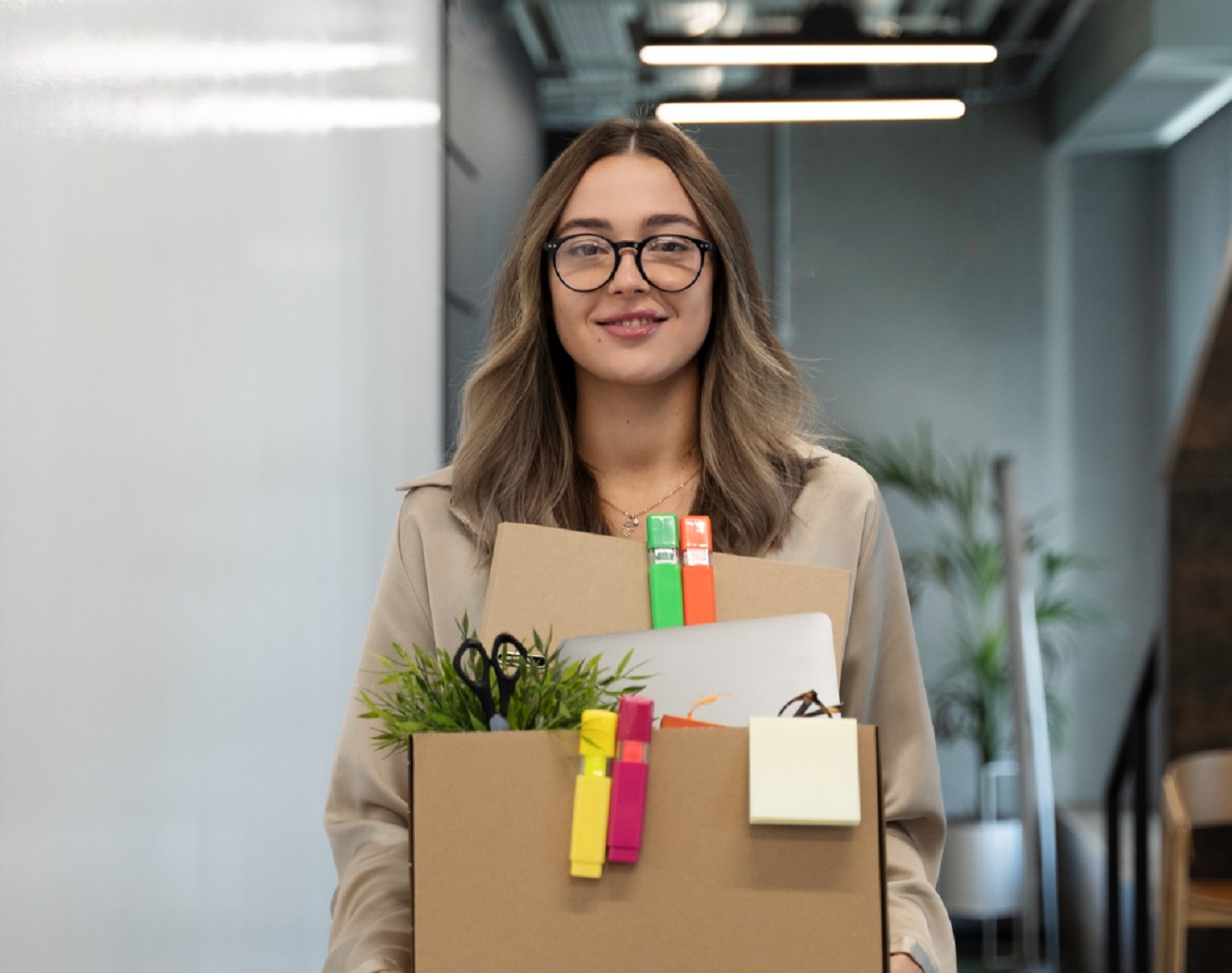 Young woman is holding a box with personal things and smiling at camera.